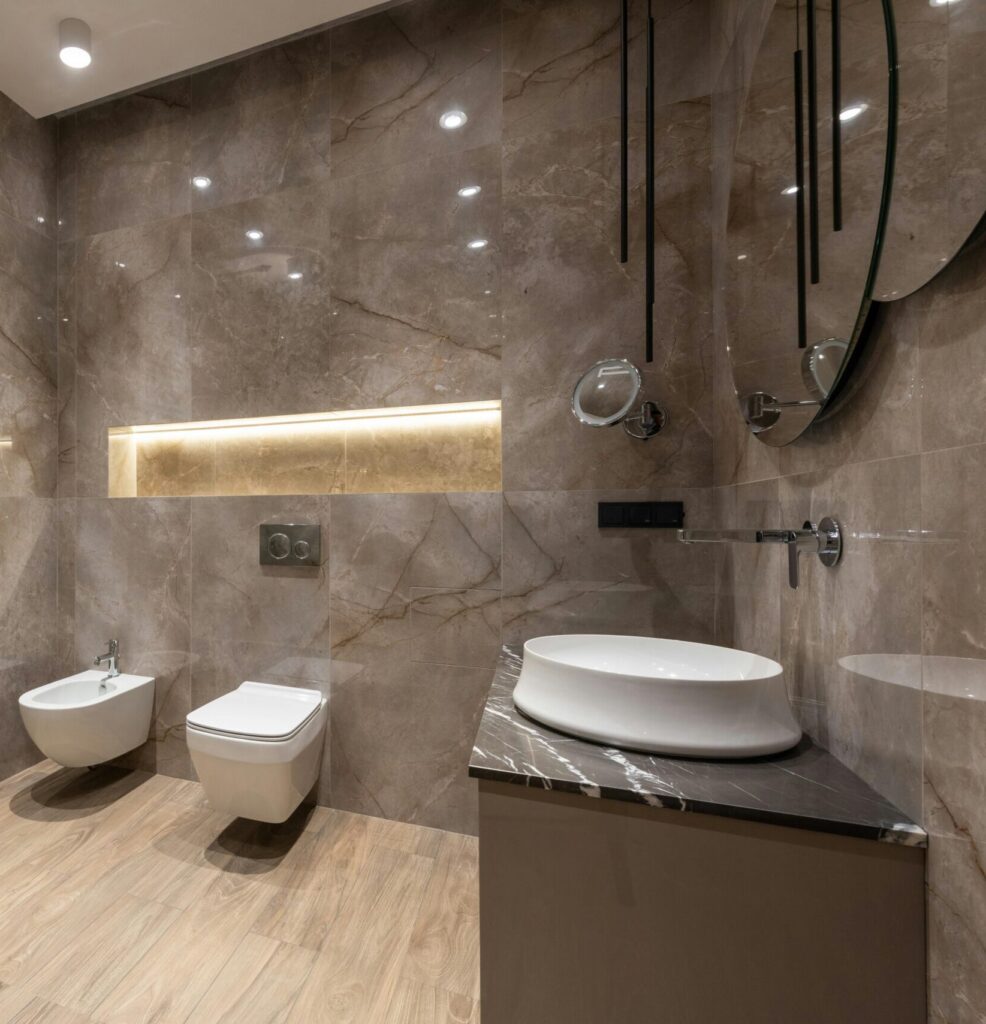 Glass shower cabin and ceramic sink with toilet bowl and bidet in spacious contemporary bathroom with shiny tiles and round mirrors